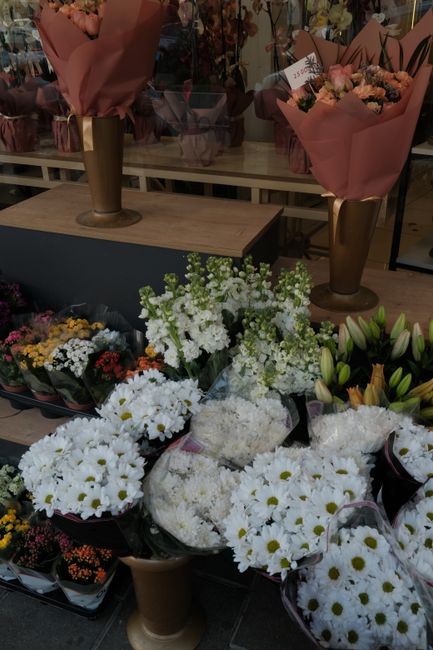 A display of fresh floral arrangements on a wooden table, featuring predominantly white flowers including daisies, chrysanthemums, and lilies, with some arrangements wrapped in pink and purple paper. 