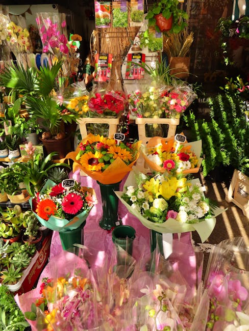 A vibrant display of fresh flower bouquets arranged on a pink table inside a florist shop. The bouquets feature a variety of colorful flowers, including bright orange, yellow, pink, and red blooms suc