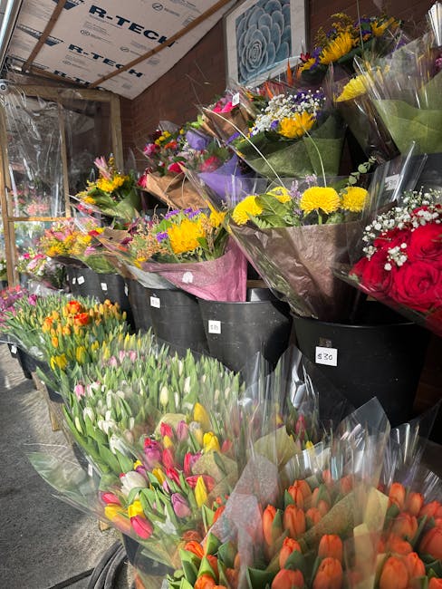 A diverse display of fresh floral bouquets arranged in black buckets at a flower shop near Willesden Junction station, Harlesden. The arrangements feature a variety of flowers including yellow sunflow