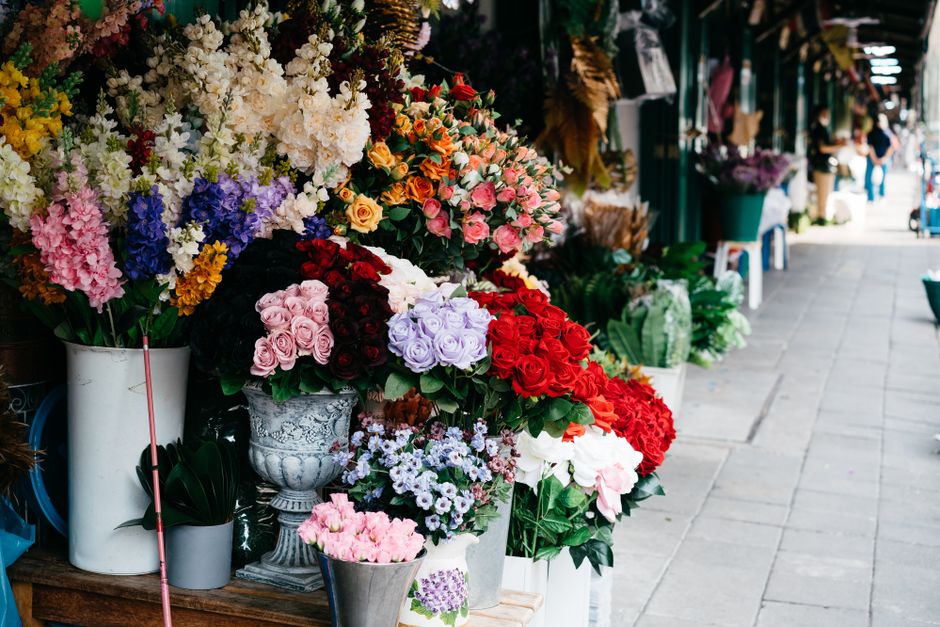 A vibrant display of fresh floral bouquets arranged in various white, silver, and glass vases on a flower shop's outdoor storefront, featuring a mix of roses in shades of pink, red, and lavender, as w