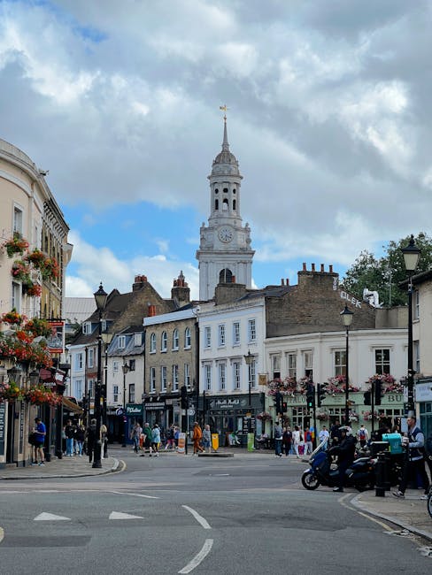 A bustling street scene in Harlesden featuring a historic clock tower with a tall, slender spire and a gold weather vane at its peak, set against a partly cloudy sky. Surrounding the tower are charmin