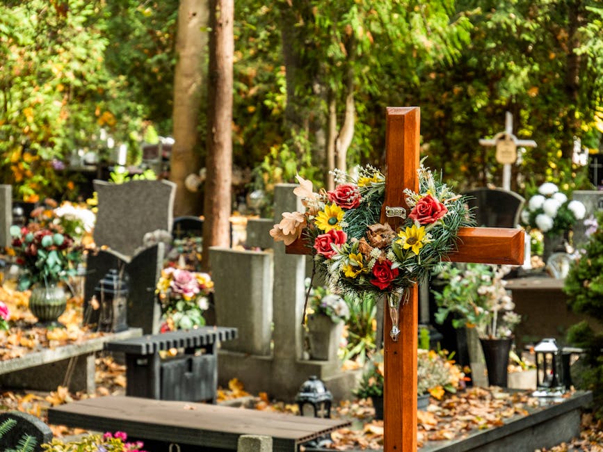 A wooden cross adorned with a floral arrangement featuring red roses, yellow daisies, and light pink blooms, surrounded by lush greenery, situated in a cemetery with gravestones and memorial plaques. 
