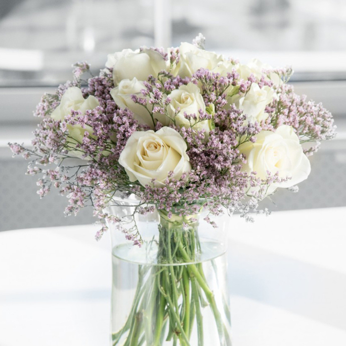 In this photograph, a refined sympathy bouquet composed of white roses and pale purple fillers is shown in close detail, as if you were standing directly in front of it in a Harlesden living room or chapel. The roses are the undeniable focal point: around a dozen large blooms, all fully open, reveal intricate spirals of petals that curl softly towards their centres. Their colour is not a stark white but a mellow, creamy ivory, with subtle shadows adding depth along the slightly ruffled edges. Between these generous roses sits an abundance of tiny lavender and dusky pink florets, likely statice or limonium, arranged in airy clusters that fill the spaces without overwhelming them. These smaller flowers create a gentle mist of pastel tones, their paper-like petals catching the light and lending a dreamy softness to the overall shape. Together, they form a rounded, full arrangement that feels both structured and natural. The stems are gathered in a tall, cylindrical glass vase with crystal-clear water, where the green stems converge in tidy lines, emphasising the freshness and care behind the design. The vase rests on a clean, white surface that reflects a subtle glow, enhancing the brightness of the roses. Behind the arrangement, the background is out of focus, rendered in bright, neutral shades that suggest an uncluttered, modern interior somewhere off Harrow Road or near Craven Park. The lighting is gentle and diffused, like a quiet mid-morning in Harlesden when the bustle of the high street is kept at a respectful distance. The bouquet conveys calm, reverence, and the quiet hope that flowers often bring to difficult days.
