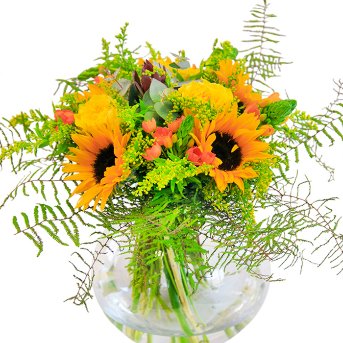 Brimming with sunlit exuberance, this striking bouquet captures the heart of a golden afternoon in Harlesden. Towering sunflowers and elegantly spiraled yellow garden roses unfurl in generous layers, their petals beaming outwards and radiating heat and happiness akin to a summer's day on Station Road. Between these vivid blossoms, emerald leaves and fine, wispy ferns stretch out, lending gentle movement and fresh greenery that calls to mind the lush, secluded corners of Harlesden's parks. Red Hypericum berries nestle among the greens, their glossy skins offering a perfect foil to the softer, velvety petals around them. The entire arrangement is cradled in a round glass vase, which reveals the organic structure of intersecting green stems and the crystal clarity of cool water upon which they're anchored. Looking closer, you'll notice the interplay of textures-the suede-like sunflowers, the tight swirls of each garden rose, the crisp snap of fresh stems. Soft scents of field flowers mingle, conjuring feelings of open air and tranquil gardens. This bouquet is the embodiment of celebration-a fitting gift for birthdays, anniversaries, or simply to bring joy into a home. Crafted by local florists who know Harlesden's rhythms and tastes, it delivers not only beauty, but also a sense of place and the happiness of shared moments.