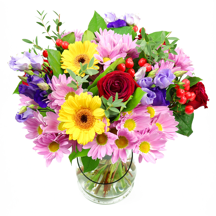 This photograph shows a richly coloured mixed bouquet displayed in a simple, elegant clear glass vase, the kind you might find gracing a dining table in a Harlesden townhouse. The arrangement is full and rounded, overflowing with blooms that almost spill beyond the edges of the frame. At the centre sit plush, deep red roses, their petals layered in tight spirals that suggest classic romance and heartfelt affection. Radiating outward, large yellow gerbera daisies with dark, warm centres create bursts of sunshine, their flat, daisy-like faces adding a graphic, modern touch that contrasts beautifully with the curves of the roses. Surrounding these key flowers, a soft ring of pink and lilac chrysanthemums and daisies adds gentle texture; each bloom is formed of many narrow petals circling around bright yellow centres, giving the impression of a soft floral cushion encircling the heart of the bouquet. Nestled among them are graceful lisianthus flowers in shades of violet and lavender, their subtly ruffled, bell-shaped petals curling at the tips, bringing an air of calm elegance to the mix. Here and there, clusters of round, glossy red hypericum berries glint like small jewels, punctuating the design and adding a touch of playful detail. Various shades of green foliage-from broad, glossy leaves to finer, lighter greenery-fill in the spaces, adding freshness, depth, and a natural, slightly untamed feel that echoes the greenery of nearby Roundwood Park or Gladstone Park. Through the transparent vase, neatly arranged stems can be seen fanning into clean water, underlining the bouquet's freshness and professional craftsmanship. The overall mood is joyful and welcoming, perfect for celebrating a birthday, offering congratulations, or simply brightening a Harlesden home with a lively, colourful presence.